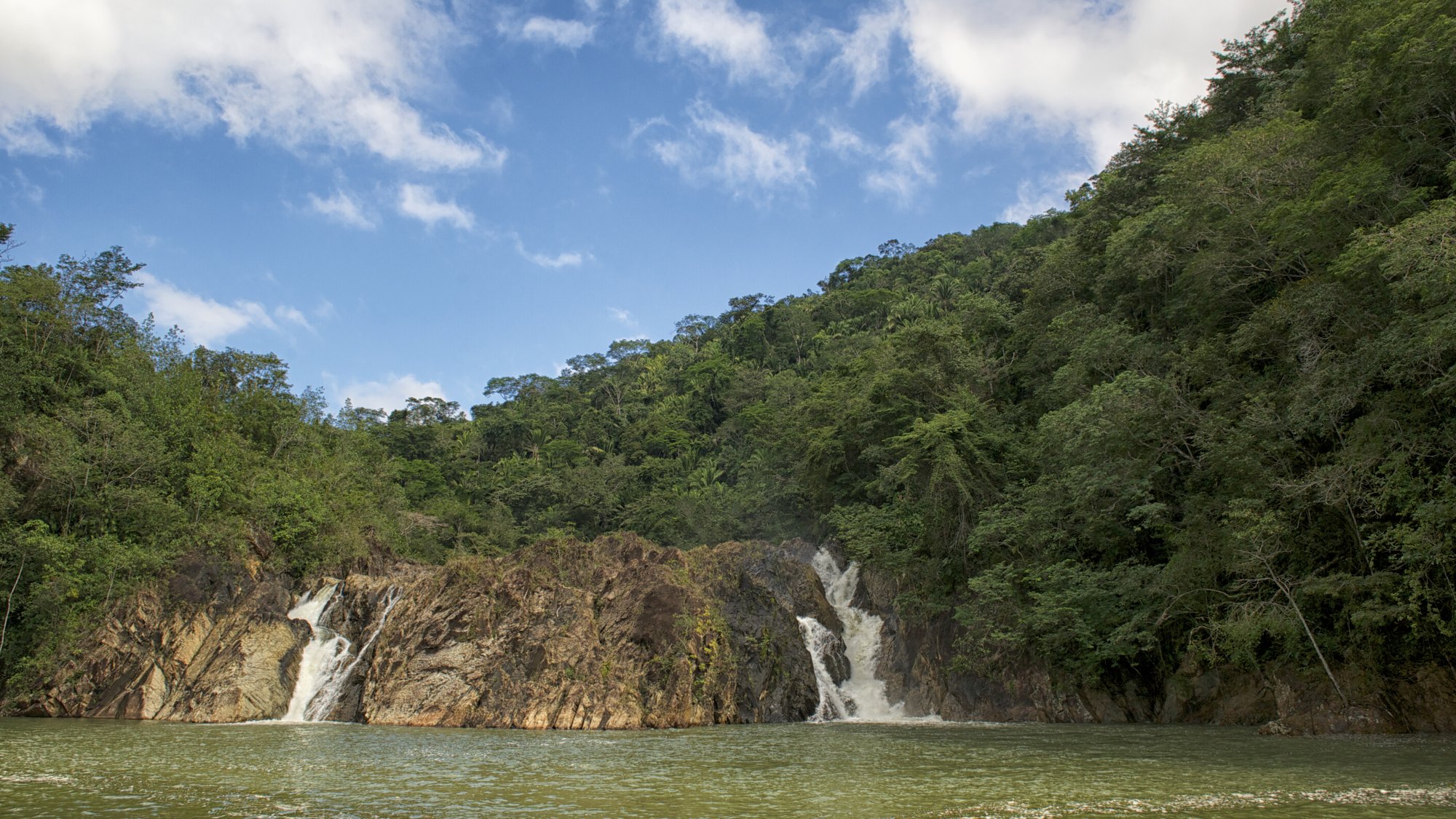 Twin waterfalls pouring into a river pool below forested hills