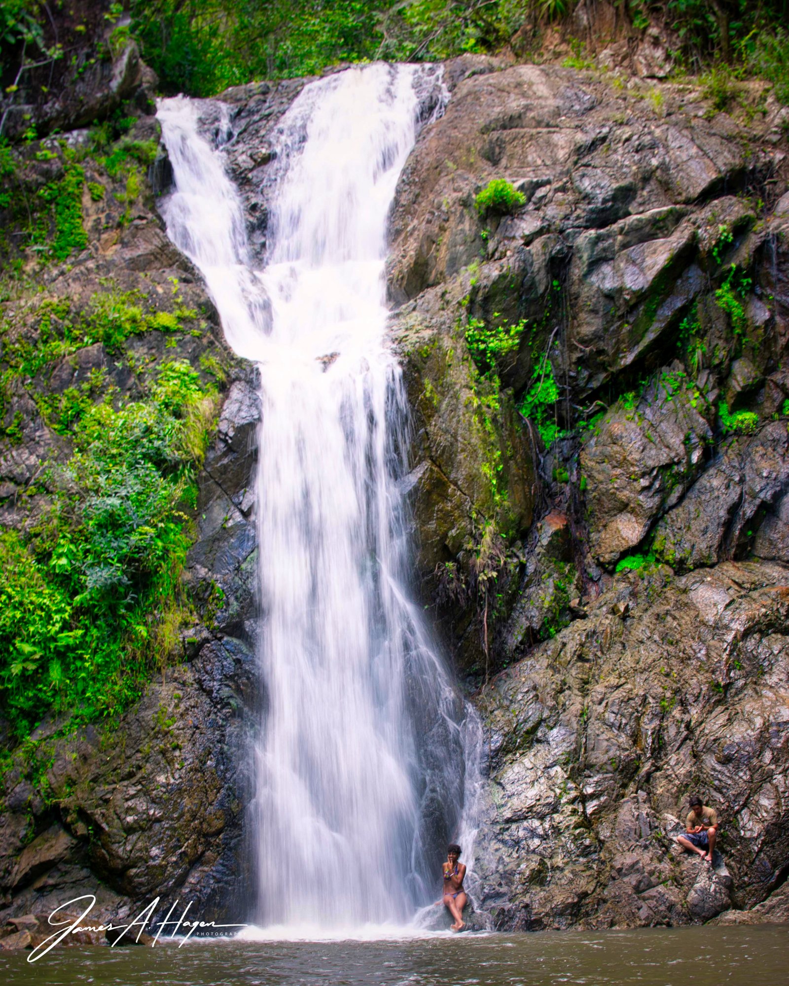 Swimmers relaxing at the base of a tall jungle waterfall